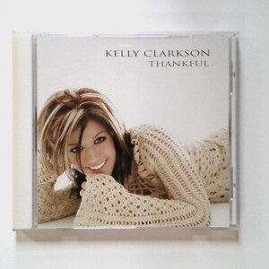 May include: A CD case featuring the album "Thankful" by Kelly Clarkson. The cover shows a smiling woman with blonde and brown hair wearing a beige crocheted top. The artist's name and album title are printed in a simple font.