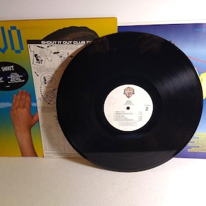 Könnte beinhalten: Eine schwarze Schallplatte mit einem weißen Etikett mit dem Bandnamen und den Titeln der Songs. Das Albumcover ist sichtbar, mit dem Wort "DEVO" in großen blauen Buchstaben und einer Handgrafik. Das Album trägt den Titel "DEVO Shout".