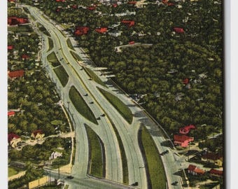 May include: Vintage postcard featuring an aerial view of the New Express Highway in Dallas, Texas. The image shows a multi-lane highway with cars, surrounded by lush green trees and buildings. The city skyline is visible in the distance.