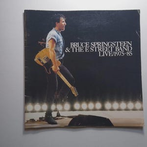 May include: Album cover for Bruce Springsteen & The E Street Band LIVE/1975-85. The image features a man holding a guitar, with text overlayed. The background is dark with stage lights.