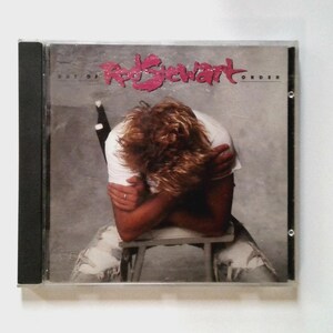 May include: CD case for Rod Stewart's "Out of Order" album. The cover art depicts a person with curly hair, wearing a white t-shirt, seated on a stool with arms crossed. The album title is in pink text.