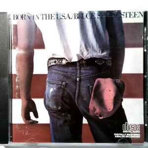 May include: CD case featuring the album "Born in the U.S.A." by Bruce Springsteen. The cover art shows a person's back in jeans and a white shirt, with an American flag background. The CD case is in a clear plastic case.