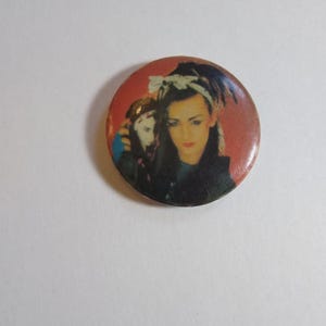 以下が含まれることがあります： ダークヘアとメイクアップの人物の肖像画が描かれた丸いボタン。ヘアバンドを着用しています。ボタンは赤い背景で、ヴィンテージな雰囲気です。