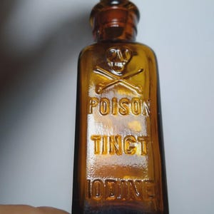 May include: A small, rectangular amber glass bottle with a cork stopper. The front features an embossed skull and crossbones symbol above the raised text POISON, TINCT, and IODINE. The glass has a textured finish and a narrow neck design.