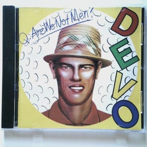 Könnte beinhalten: Eine Musik-CD-Hülle mit dem Albumcover von Devos Q: Are We Not Men? Das Kunstwerk zeigt eine stilisierte Illustration einer Person mit einem karierten Fischerhut vor einem gelben kreisförmigen Hintergrund mit schwarzen Halbmonden und dem Wort DEVO.