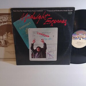 May include: A vinyl record album cover for the film "Midnight Express" with a black background and red lettering. The cover features a passport and a photo of a person with their hands raised. A second album cover is visible to the left.