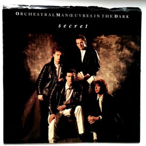 May include: Album cover for Orchestral Manoeuvres in the Dark's "Secret" album. The image shows four men in black jackets and white shirts against a dark backdrop. The band's name and album title are at the top.