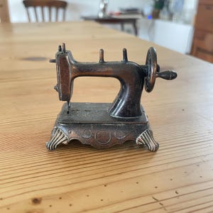 May include: A miniature sewing machine figurine crafted from dark bronze-toned metal. The object features a hand crank on the side, vertical spool pins, and a rectangular base with decorative scrollwork and silver-toned feet.