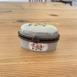 May include: An oval porcelain trinket box featuring a white base with a green lattice pattern and gold accents. The lid and front panel display hand-painted floral motifs in pink, yellow, and purple. It has a hinged metal closure with a flower clasp.