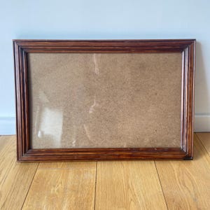 May include: A rectangular picture frame with a dark brown wood grain finish. The frame features a multi-tiered, stepped molding profile and contains a flat, light brown fiberboard backing panel set within the inner recessed edge.
