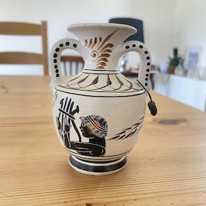 Puede incluir: Un pequeño jarrón ánfora de cerámica con base de color crema y detalles pintados en negro. El diseño presenta una figura tocando una lira, patrones de hojas decorativas alrededor del cuello y dos asas con puntos negros. Una pequeña etiqueta negra cuelga de un asa.