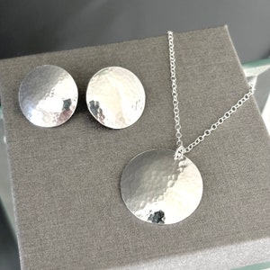 May include: A silver necklace and earrings set with hammered round pendant and matching earrings. The jewelry is on a gray background.