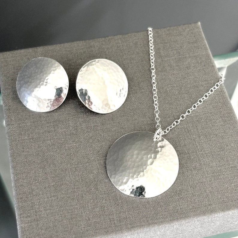 May include: A silver necklace and earrings set with hammered round pendant and matching earrings. The jewelry is on a gray background.