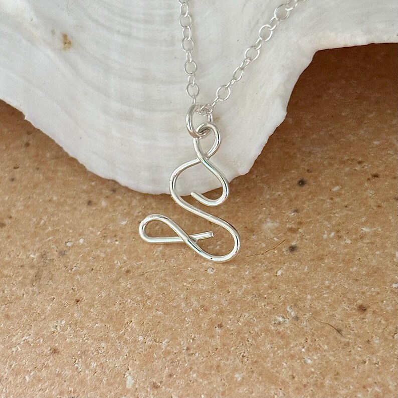 May include: A silver necklace featuring a delicate, abstract pendant shaped like a figure in a seated pose. The pendant is crafted from thin, twisted wire.