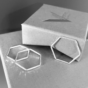 May include: Two silver hexagon rings on a grey textured surface. The rings are stacked on top of each other. The surface is a box with a logo on the top.
