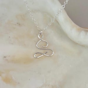 May include: A silver necklace featuring a delicate, abstract pendant shaped like a stylised figure in a seated position. The pendant is crafted from thin, twisted wire.