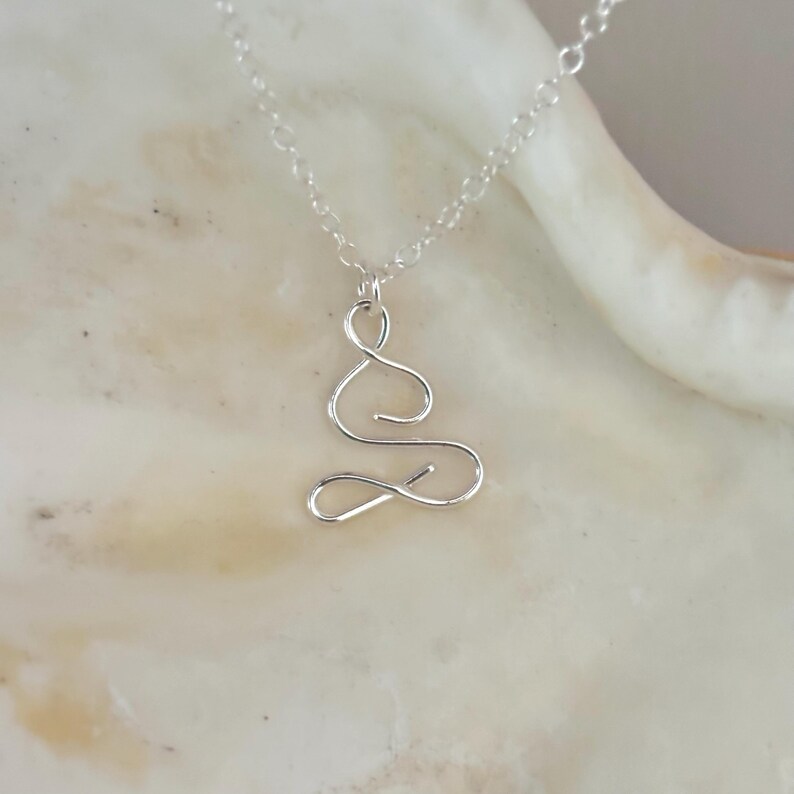 May include: A silver necklace featuring a delicate, abstract pendant shaped like a stylised figure in a seated position. The pendant is crafted from thin, twisted wire.