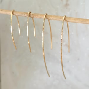 May include: Three pairs of gold-toned earrings with a curved, hammered design. The earrings are hanging from a wooden stick.
