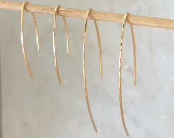 May include: Three pairs of gold-toned earrings with a curved, hammered design. The earrings are hanging from a wooden stick.