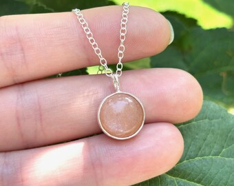May include: A silver chain necklace featuring a round, smooth, peach-coloured stone pendant set in a simple silver bezel. The pendant hangs from a delicate, thin silver link chain, showcasing a polished, circular gemstone with a soft, warm hue.