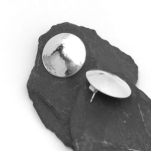 May include: A pair of silver hammered disc earrings. The earrings are round and have a textured surface. They are shown on a dark gray stone background.