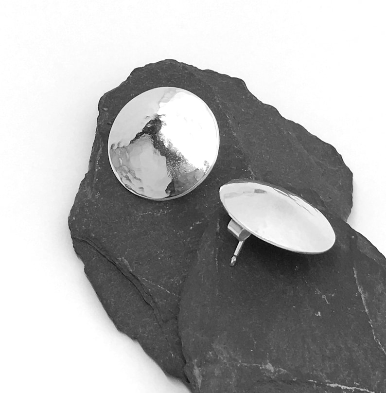 May include: A pair of silver hammered disc earrings. The earrings are round and have a textured surface. They are shown on a dark gray stone background.