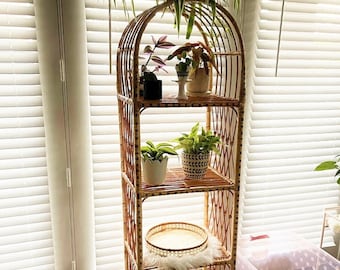 May include: A four-tiered arched rattan shelf unit with a woven design. The shelf is filled with potted plants, including a trailing plant, a fern, and a succulent. The shelf is standing on a wood floor in front of a window with white blinds.