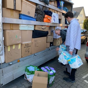 May include: A moving truck is loaded with boxes, some of which are labeled. Two people are standing near the truck, one is loading boxes and the other is holding a package of diapers.