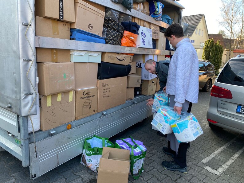May include: A moving truck is loaded with boxes, some of which are labeled. Two people are standing near the truck, one is loading boxes and the other is holding a package of diapers.