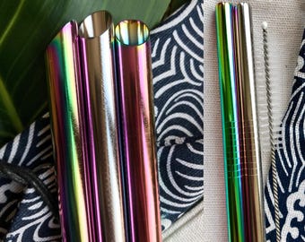 May include: Close-up of several iridescent metal straws, reflecting a spectrum of colors. The straws are cylindrical with angled tips. A cleaning brush is visible. The straws are placed on a patterned fabric with a leaf in the background.