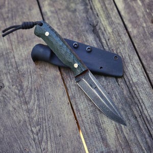 Puede incluir: Un cuchillo pequeño de hoja fija con un mango texturizado verde oscuro y una hoja plateada. El cuchillo descansa sobre una superficie de madera junto a una funda negra. Un cordón negro está unido al mango.