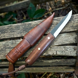 Könnte beinhalten: Ein Messer mit feststehender Klinge, dunklem Holzgriff, zwei silbernen Nieten und einer braunen Paracord-Fangschnur, dazu eine passende braune Lederscheide mit eingeprägtem Sonnenmotiv. Die Klinge hat ein dunkles Finish und eine polierte Schneide.
