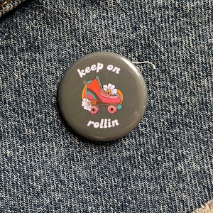 May include: A gray button with a pink and blue roller skate with white flowers and the text "keep on rollin'" in white.