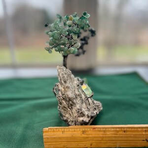 May include: A green gemstone wire wrapped tree sculpture on a rock base. The tree is made of wire and green gemstones. The base is made of a rough, gray rock.