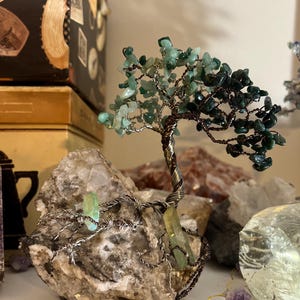 May include: A green and brown wire-wrapped crystal tree with a natural stone base. The tree is made of small green and brown crystals and is wrapped in copper wire. The base is a natural stone with a rough texture.
