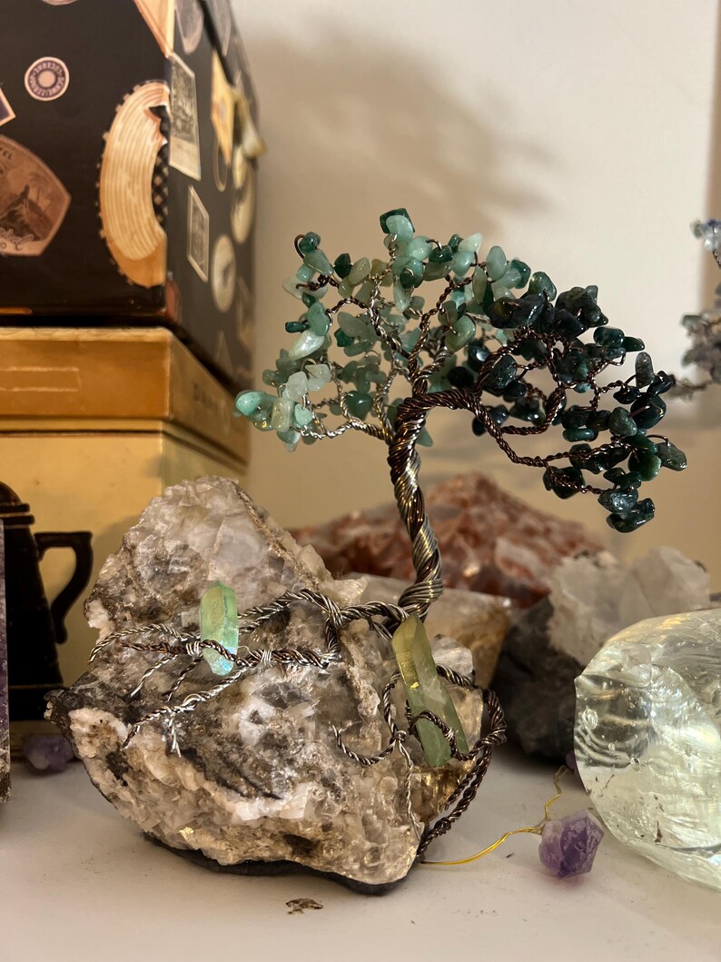 May include: A green and brown wire-wrapped crystal tree with a natural stone base. The tree is made of small green and brown crystals and is wrapped in copper wire. The base is a natural stone with a rough texture.
