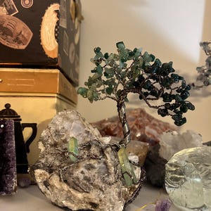 May include: A wire-wrapped tree sculpture with green gemstone leaves and a natural crystal base. The tree is made with copper wire and the leaves are made with green aventurine gemstones. The base is a natural crystal with a rough texture.
