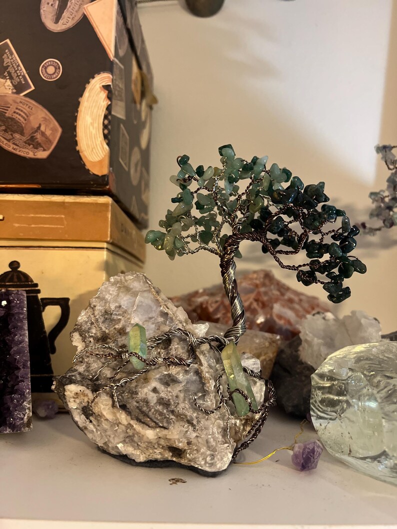 May include: A wire-wrapped tree sculpture with green gemstone leaves and a natural crystal base. The tree is made with copper wire and the leaves are made with green aventurine gemstones. The base is a natural crystal with a rough texture.