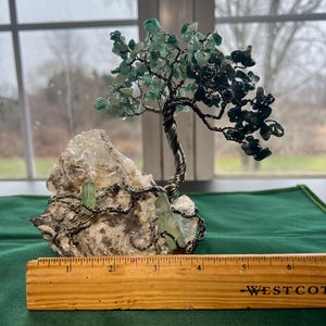 May include: A wire-wrapped green gemstone tree sculpture with a base of white crystal rock. The tree is approximately 4 inches tall and is displayed on a wooden ruler.