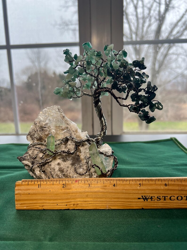 May include: A wire-wrapped green gemstone tree sculpture with a base of white crystal rock. The tree is approximately 4 inches tall and is displayed on a wooden ruler.