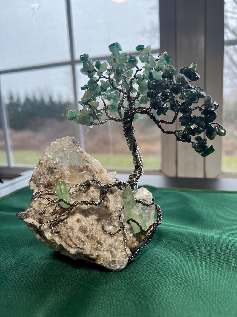 May include: A wire-wrapped crystal tree with green stones and a white crystal base. The tree is on a green surface.