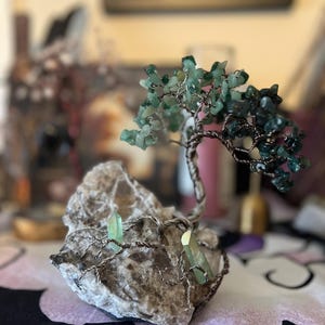 May include: A wire-wrapped tree sculpture with green gemstone leaves and branches. The tree is rooted in a gray rock with two green gemstone crystals.