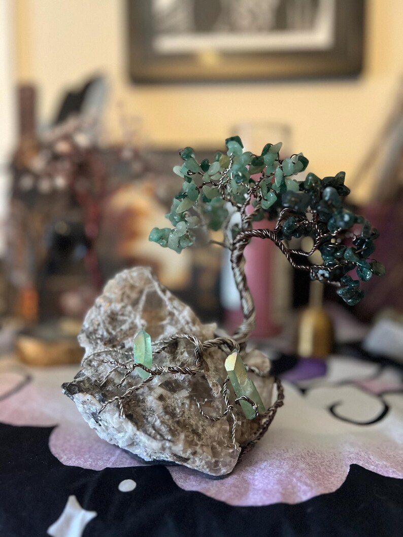 May include: A wire-wrapped tree sculpture with green gemstone leaves and branches. The tree is rooted in a gray rock with two green gemstone crystals.