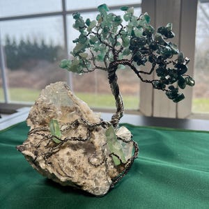 May include: A wire-wrapped tree sculpture with green gemstone leaves and a white crystal base. The tree is made of wire and has a natural, organic look. The base is a rough, white crystal with a few green crystals embedded in it.