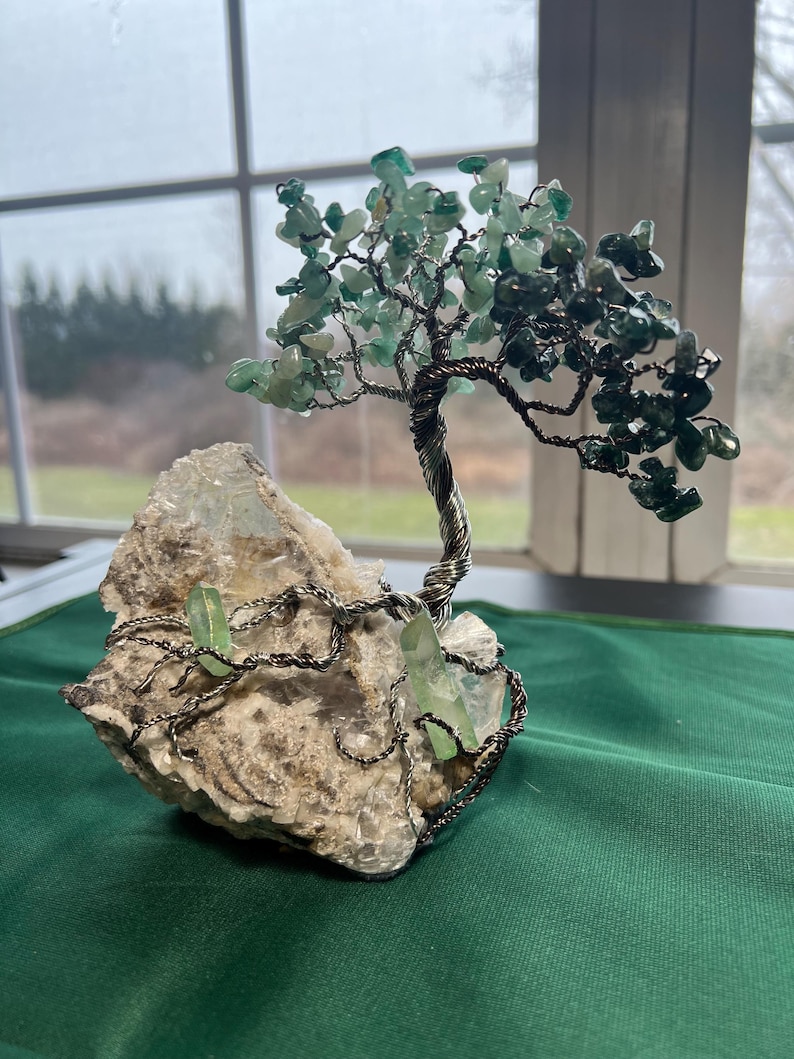 May include: A wire-wrapped tree sculpture with green gemstone leaves and a white crystal base. The tree is made of wire and has a natural, organic look. The base is a rough, white crystal with a few green crystals embedded in it.