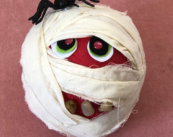 May include: A round red object wrapped in off-white fabric strips resembling a mummy. It features two large eyes with green and black pupils, three small light-colored stone-like teeth at the bottom, and a black plastic spider on the top.