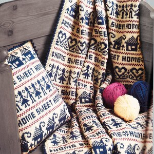 May include: A cream-coloured knitted blanket and cushion set with a navy blue and burgundy pattern. The design includes houses, hearts, and people, with the words "HOME SWEET HOME". The set is draped over a wooden bench.
