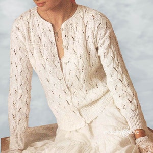 May include: Cream-coloured, open-weave cardigan with a delicate, textured pattern. The long-sleeved sweater features a button-down front and is paired with a matching skirt. A straw hat with a pink ribbon adds a touch of elegance.