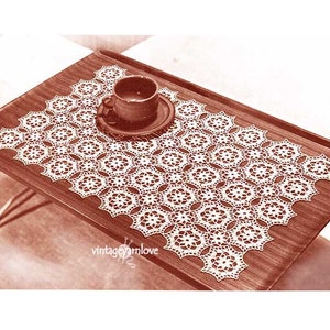 May include: A white crocheted table runner with a delicate floral pattern. The runner is laid out on a wooden table with a cup and saucer on top.
