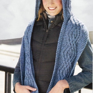 May include: A blue knitted hooded scarf with cable knit detail and pockets. The scarf is worn by a person with a black vest and blue jeans.